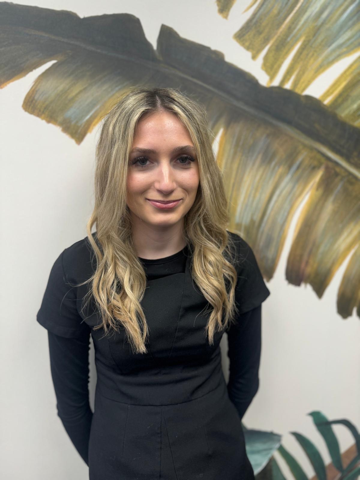 Salon apprentice Shannon stood in front of floral wallpaper in black top with long sleeves